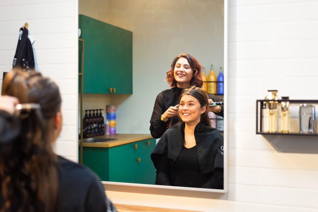 A smiling female hairdresser with auburn hair styles a smiling customer's hair with a straightener, seen through a large rectangular mirror reflection inside a modern salon with emerald green cabinets.