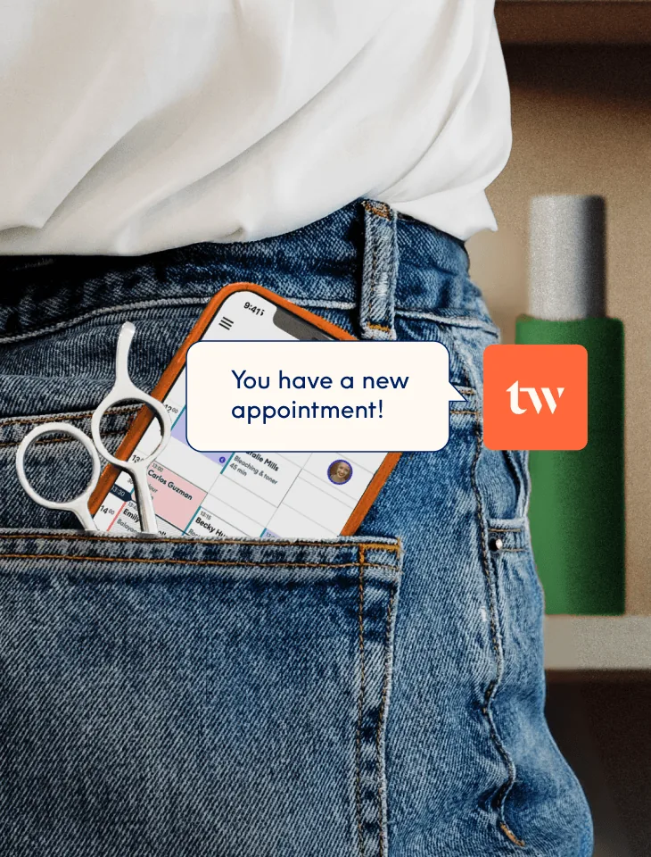 A smartphone and hairdressing scissors tucked into a jeans back pocket, the screen showing the Treatwell app's booking calendar with a "new appointment" notification.